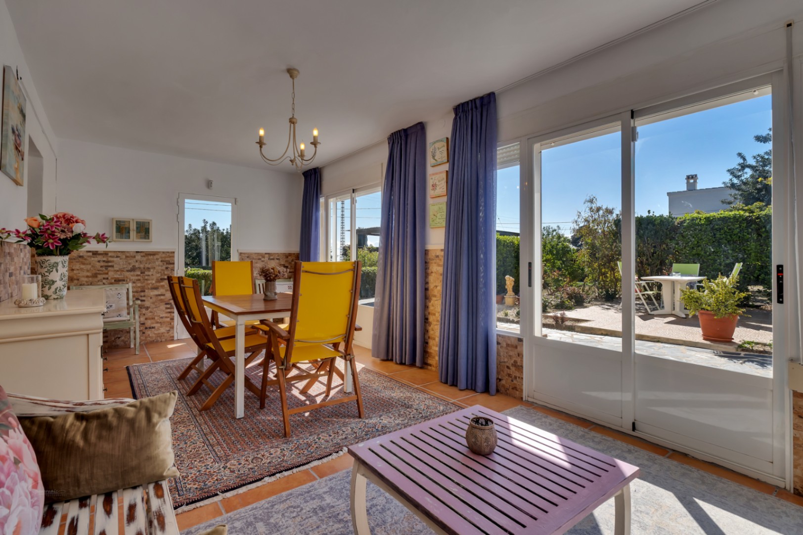 Bright dining and living area featuring yellow chairs, stone-effect wall accents, and large sliding glass doors opening onto the sunny garden terrace.