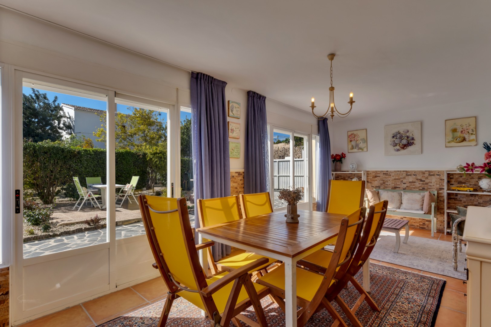 Bright dining area featuring a wooden table with yellow chairs, terracotta flooring, and large sliding doors opening onto the sunlit garden terrace.