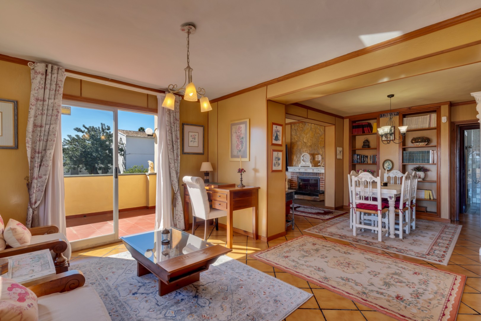 Bright living and dining area featuring a wooden desk, tiled flooring with rugs, and terrace access. The background shows a dining set and built-in bookshelves.
