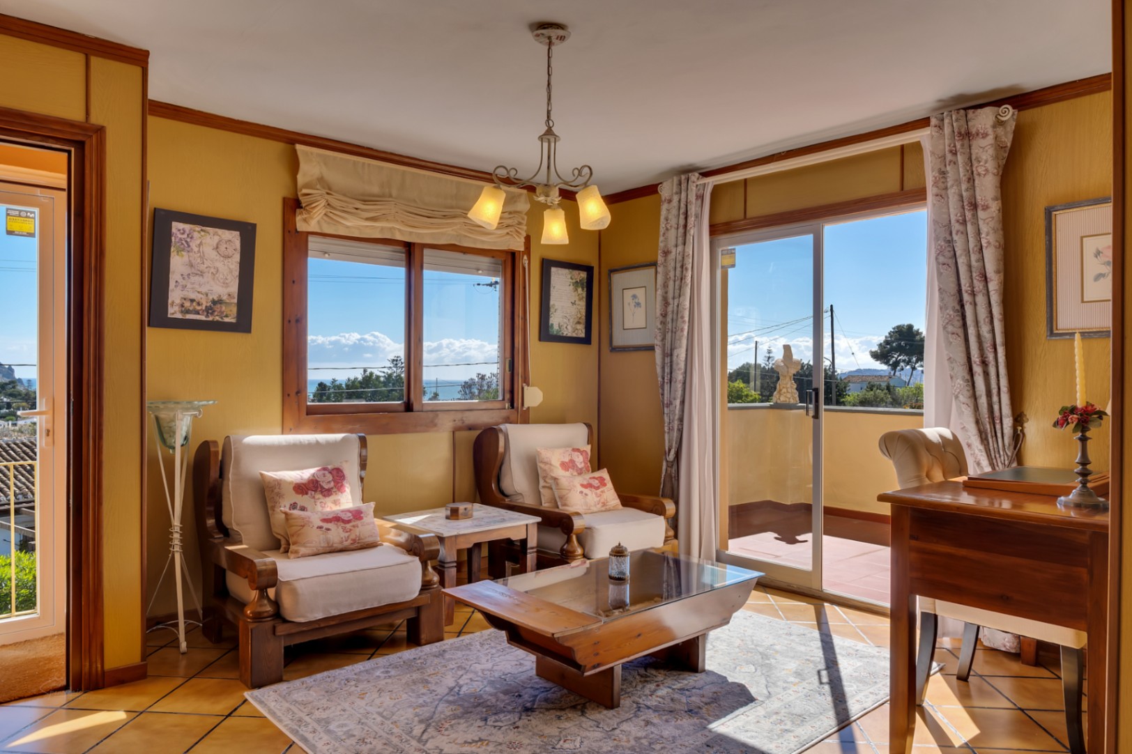 Bright living area featuring wooden armchairs, a glass-top coffee table, and large sliding doors opening onto a terrace with Mediterranean sea views.