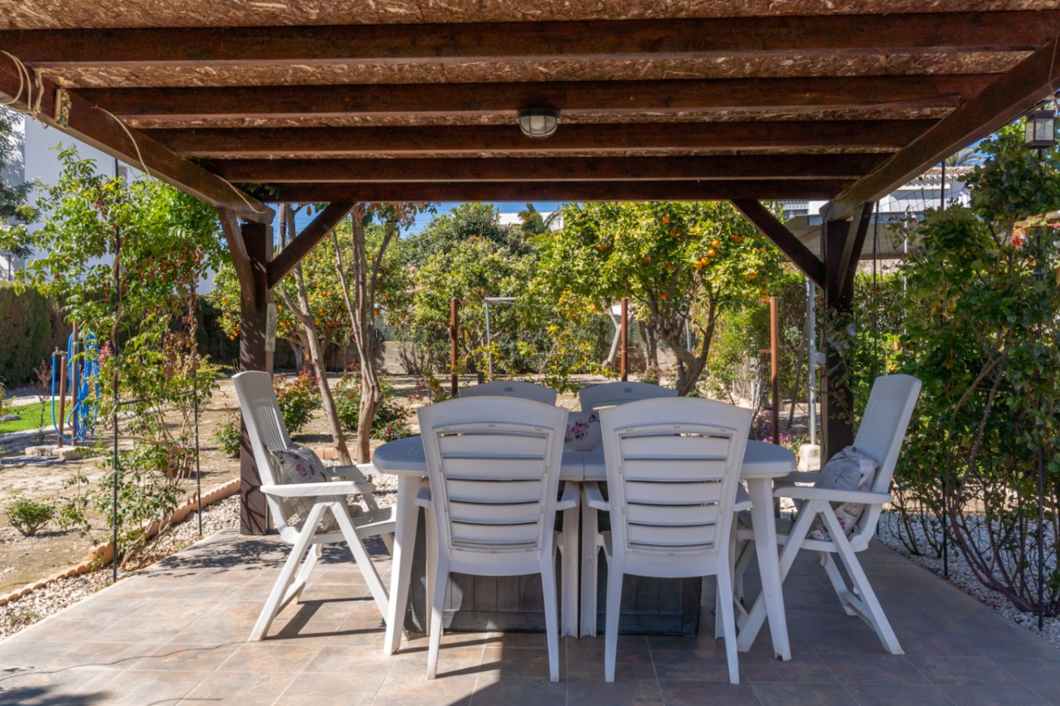 Covered outdoor dining area featuring a wooden pergola and white furniture, overlooking a garden with citrus trees.