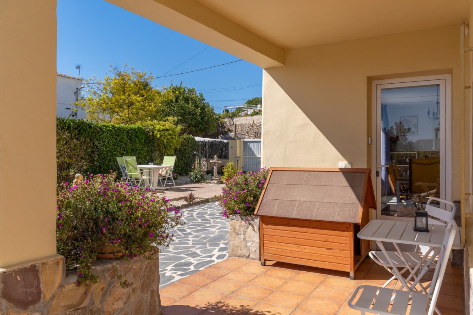 Covered porch with terracotta tiles overlooking a garden with stone paths, mature hedges, and an outdoor dining area under the Mediterranean sun.
