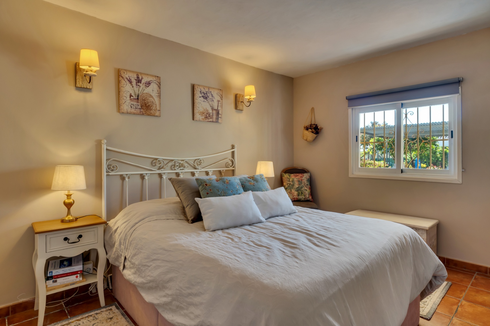 Cozy bedroom featuring a white wrought iron headboard, terracotta tile flooring, and a window with decorative security bars overlooking the garden.