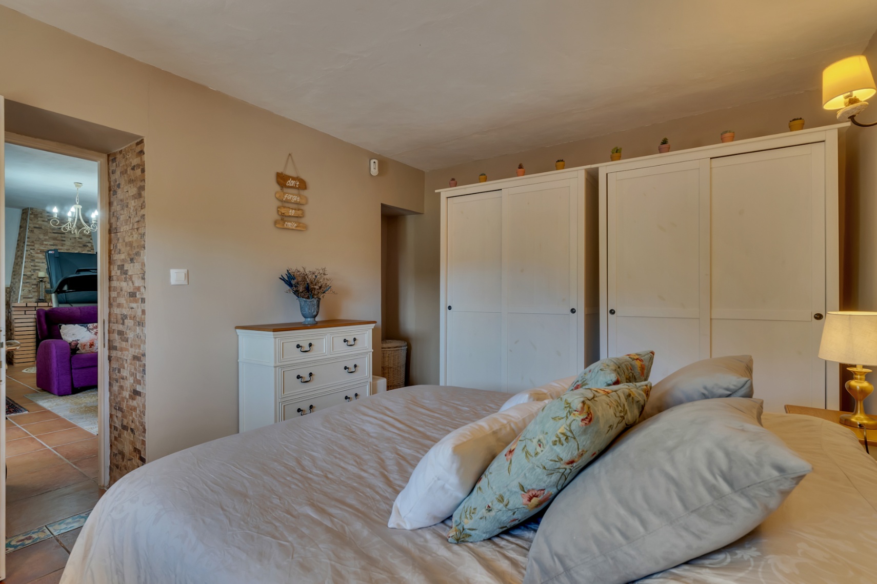 Cozy bedroom with white wooden wardrobes, a matching dresser, and earth-toned walls. A natural stone accent frames the doorway leading to the adjacent living room.