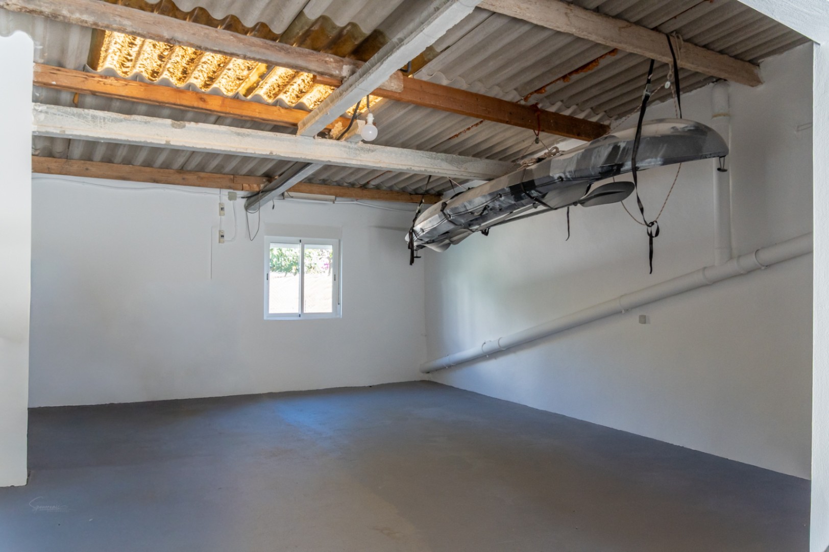 Enclosed garage with grey flooring and wooden beams, featuring a suspended kayak storage system and a corrugated roof with natural light.