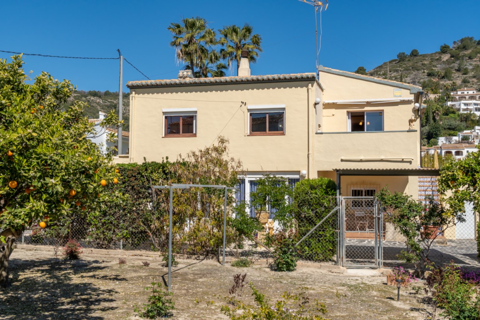 Exterior facade of the two-story villa featuring cream-colored walls, citrus trees, and palm trees set against a Mediterranean hillside.