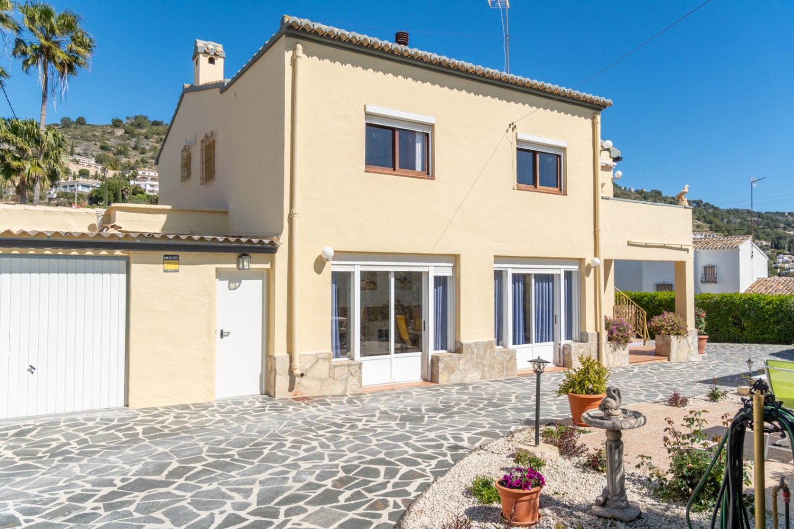 Exterior view of the villa featuring a cream facade with stone accents and a spacious paved patio. Includes a private garage and large white-framed windows.