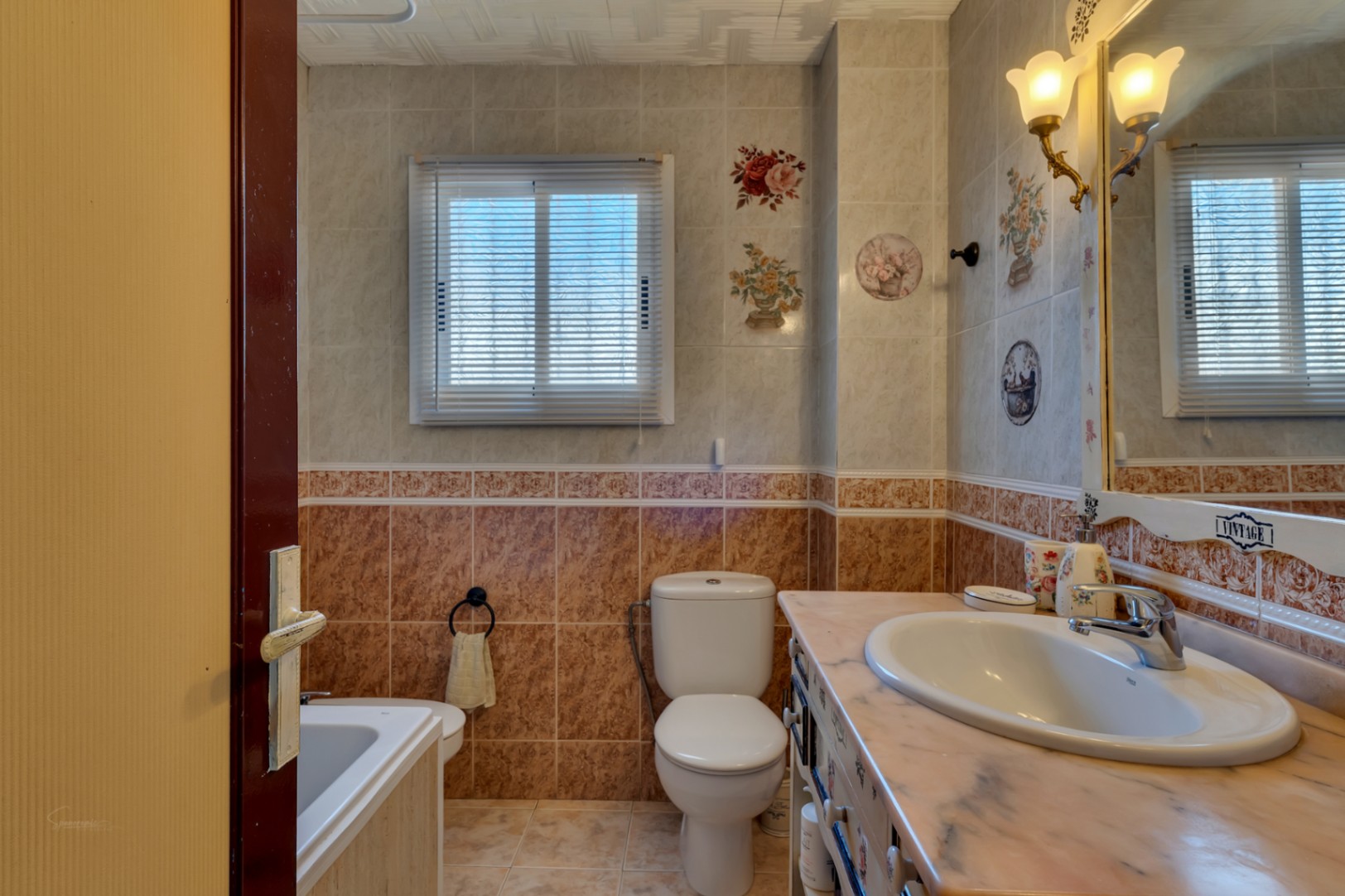 Full bathroom featuring decorative floral tiles, a marble vanity top, classic wall sconces over the mirror, and a window with integrated blinds.