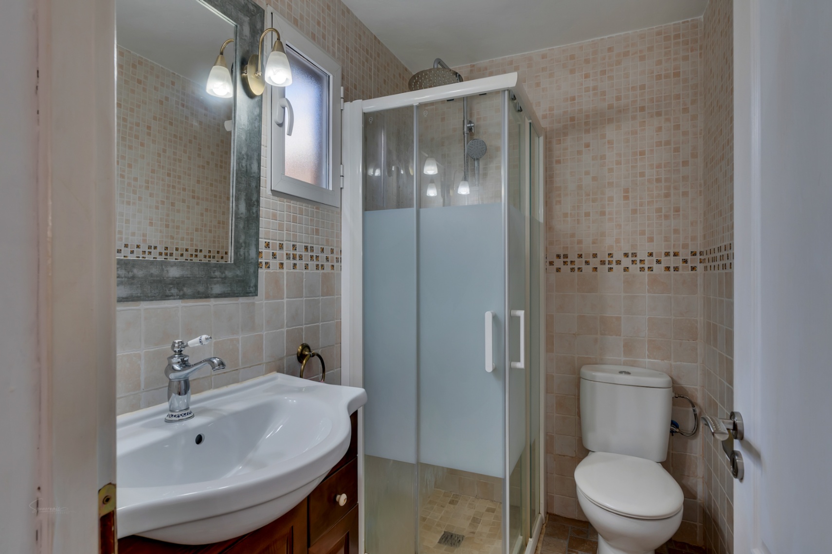 Functional bathroom featuring beige tiled walls, a glass-enclosed shower, a white ceramic sink with wooden vanity, and a mirror with traditional wall sconces.
