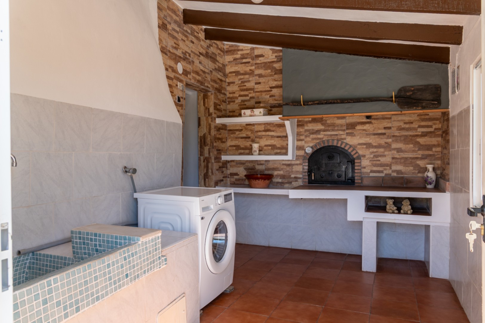Functional laundry and rustic kitchen area featuring a blue mosaic sink, washing machine, and traditional wood-fired oven set against exposed dark wood ceiling beams.