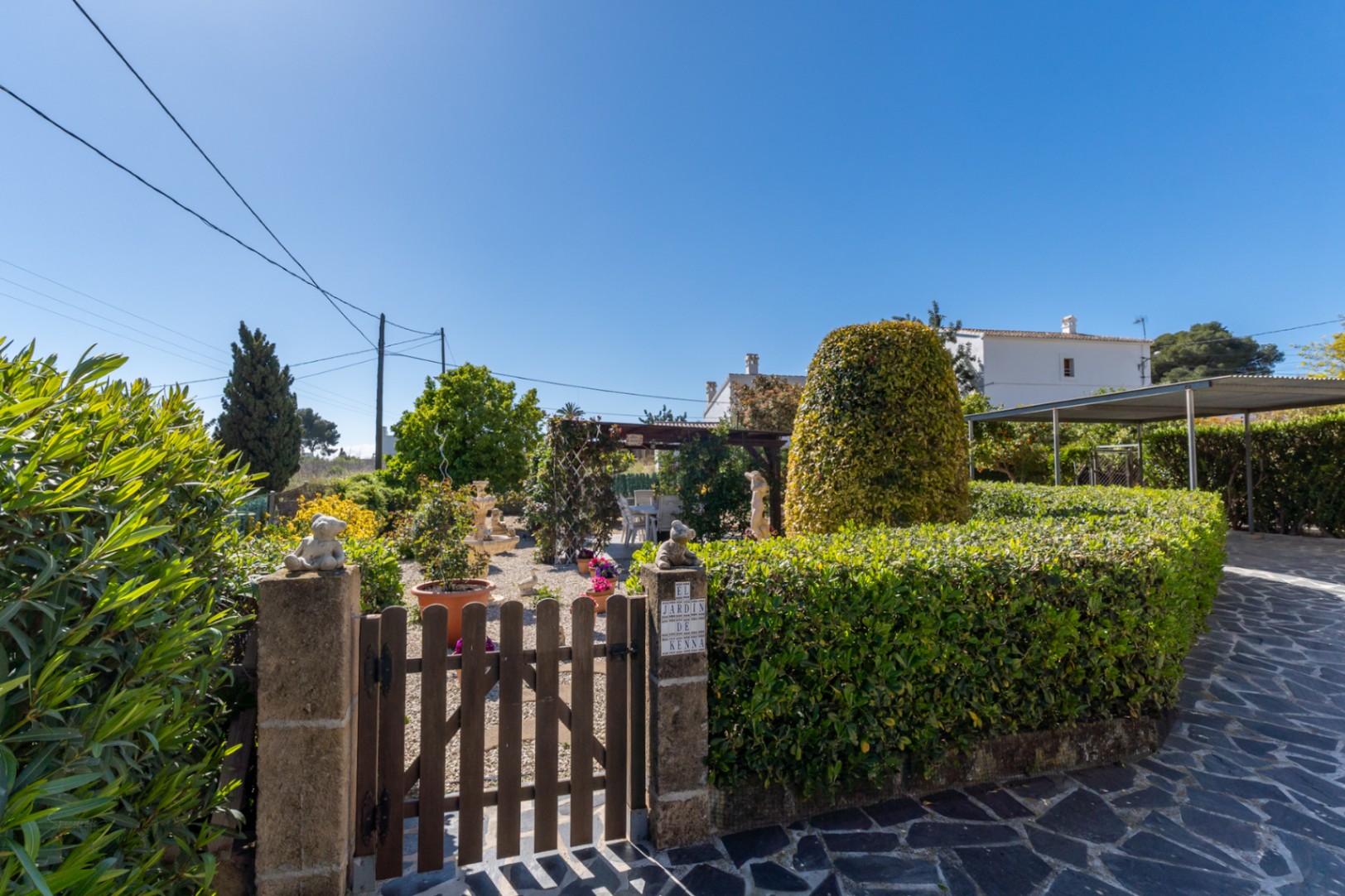 Garden entrance featuring a wooden gate, stone pillars, and manicured hedges. The gravel path leads to a pergola, with a carport and stone driveway to the side.