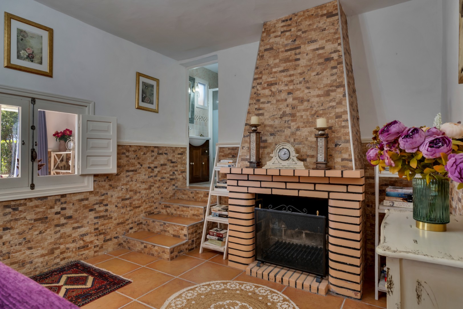 Living area featuring a brick fireplace with stone cladding, terracotta tile flooring, and steps leading to a bathroom. Traditional white shutters and rustic decor define the space.