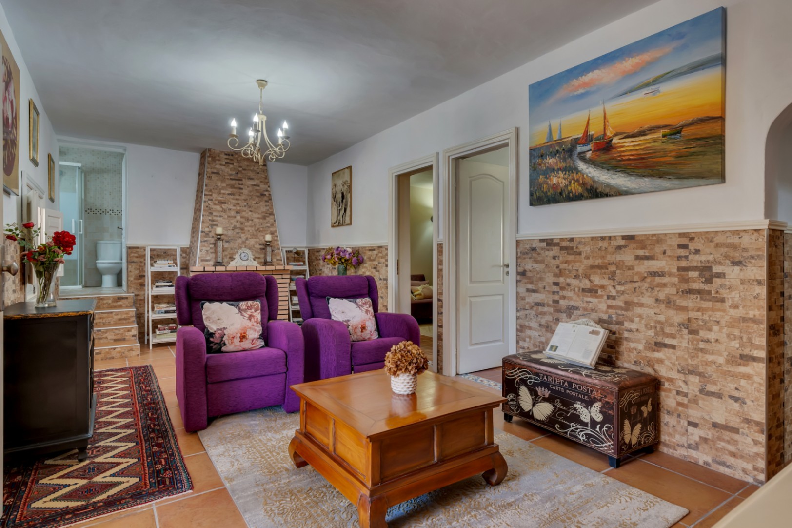 Living area featuring a stone-clad fireplace, purple armchairs, and ceramic tile flooring. The space connects to adjacent rooms and a visible bathroom.