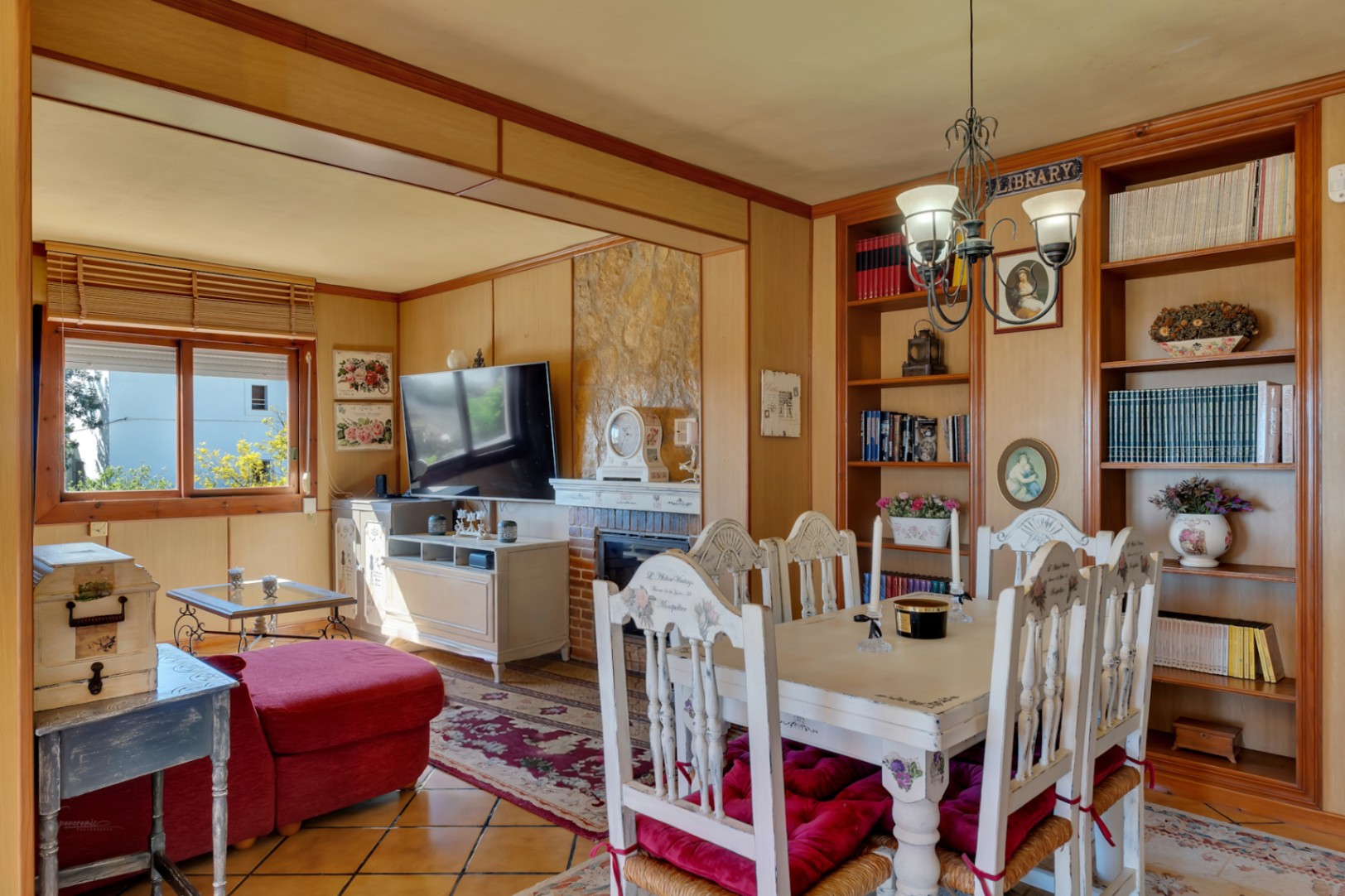 Living-dining area featuring a stone fireplace, wood-paneled walls, and built-in bookshelves. The space includes rustic furniture and traditional ceramic tile flooring.