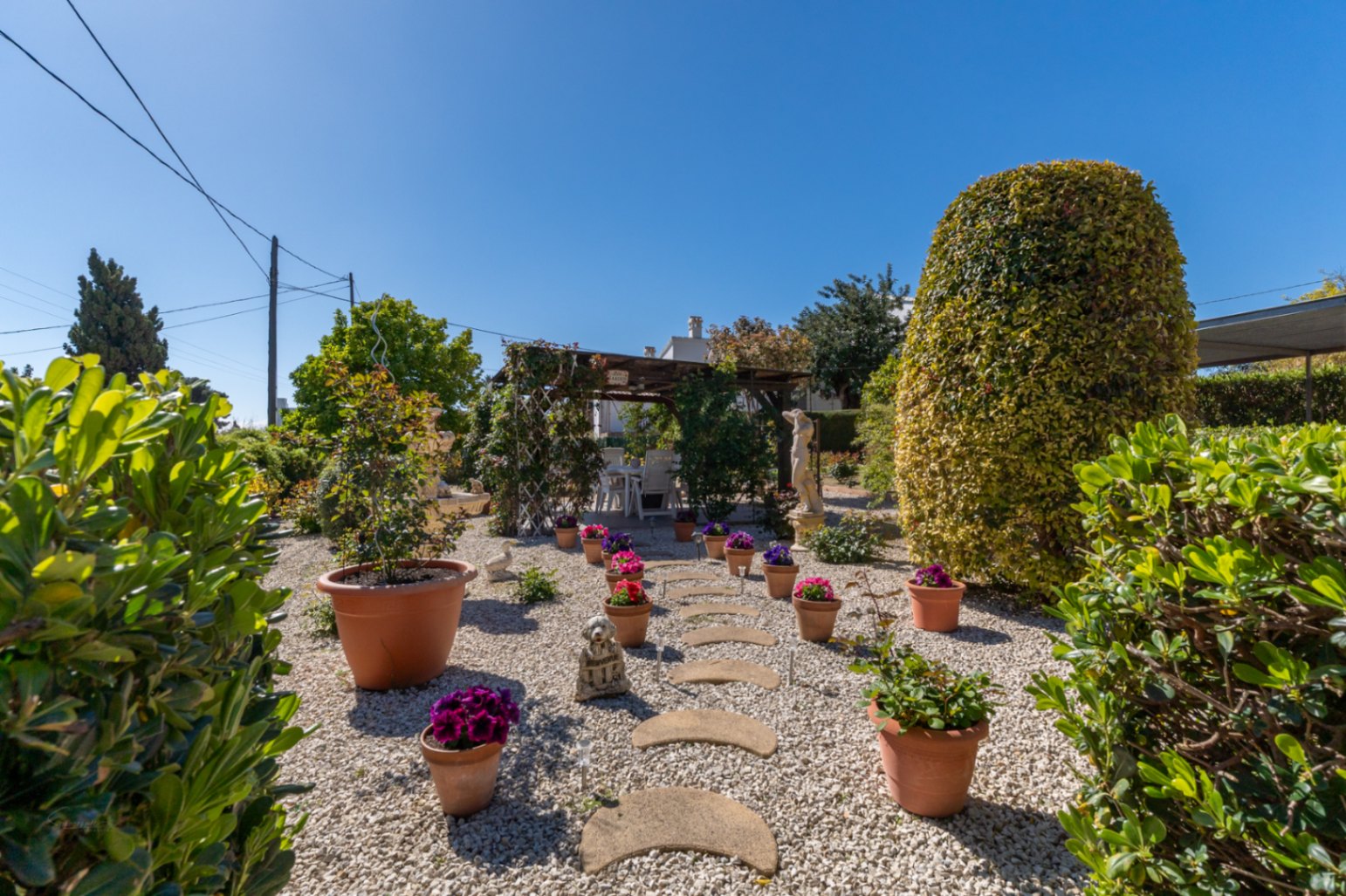 Mediterranean garden featuring white gravel, a stone stepping path, and terracotta pots. A wooden pergola houses an outdoor dining area amidst manicured greenery.