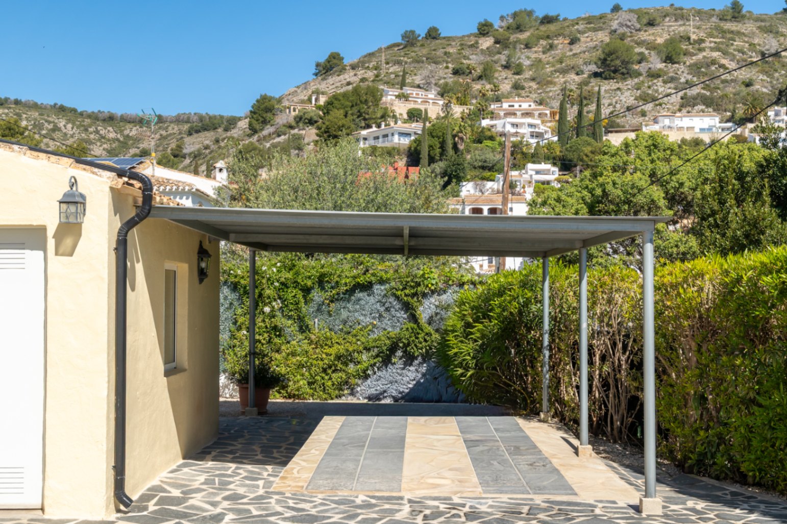 Metal carport with a flat roof and stone-paved driveway, located beside the main building with views of the surrounding Mediterranean hillside.