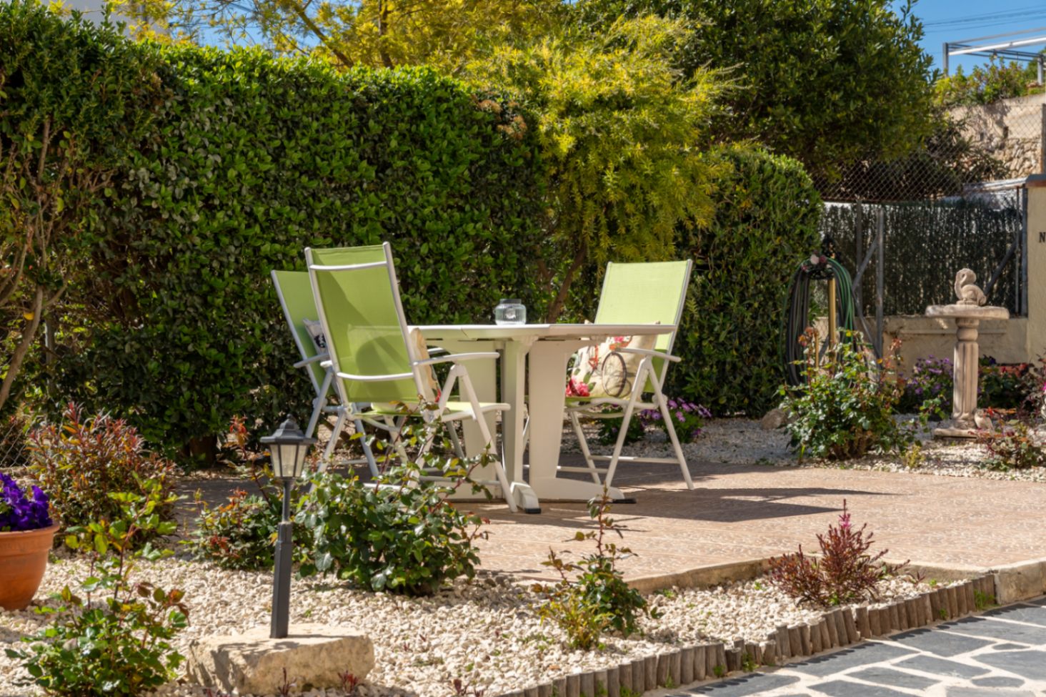 Outdoor seating area featuring a white table and lime green chairs, set on a tiled patio surrounded by decorative gravel and lush privacy hedges.