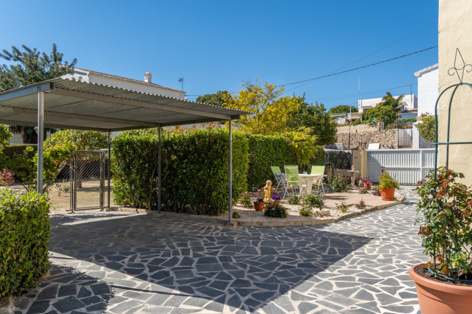 Property entrance featuring an irregular stone-paved driveway, a metal carport, and a landscaped garden area with mature hedges and seating.