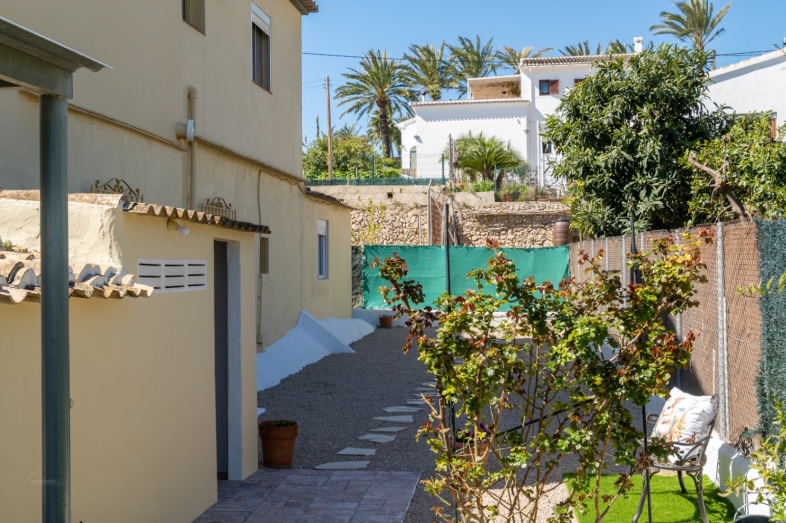 Side exterior area featuring a gravel path with stepping stones, Mediterranean shrubbery, and a functional outbuilding under bright natural light.