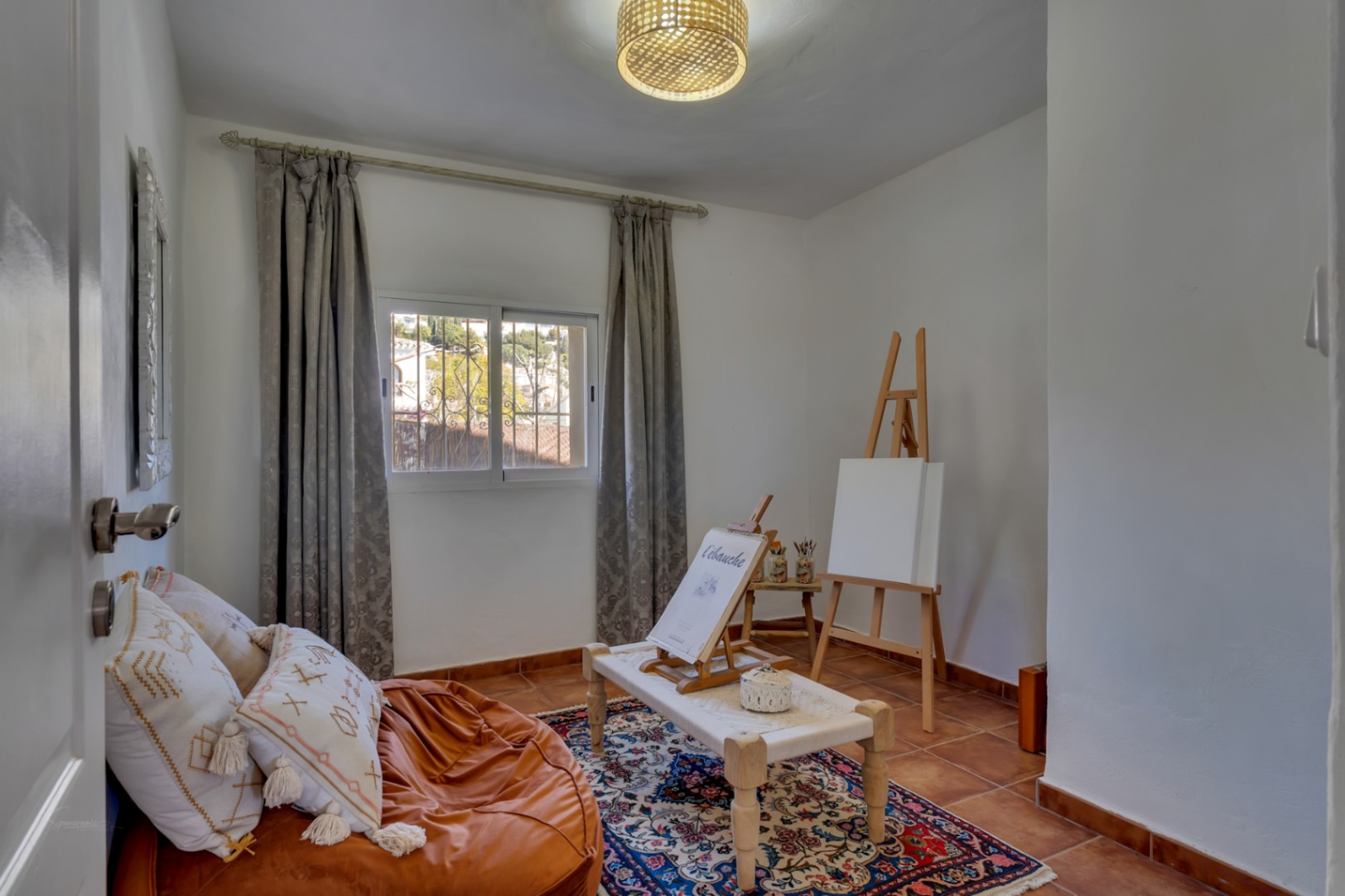Versatile creative studio featuring traditional terracotta tile flooring, a wooden easel, natural window light, and a relaxed seating area with a bean bag.
