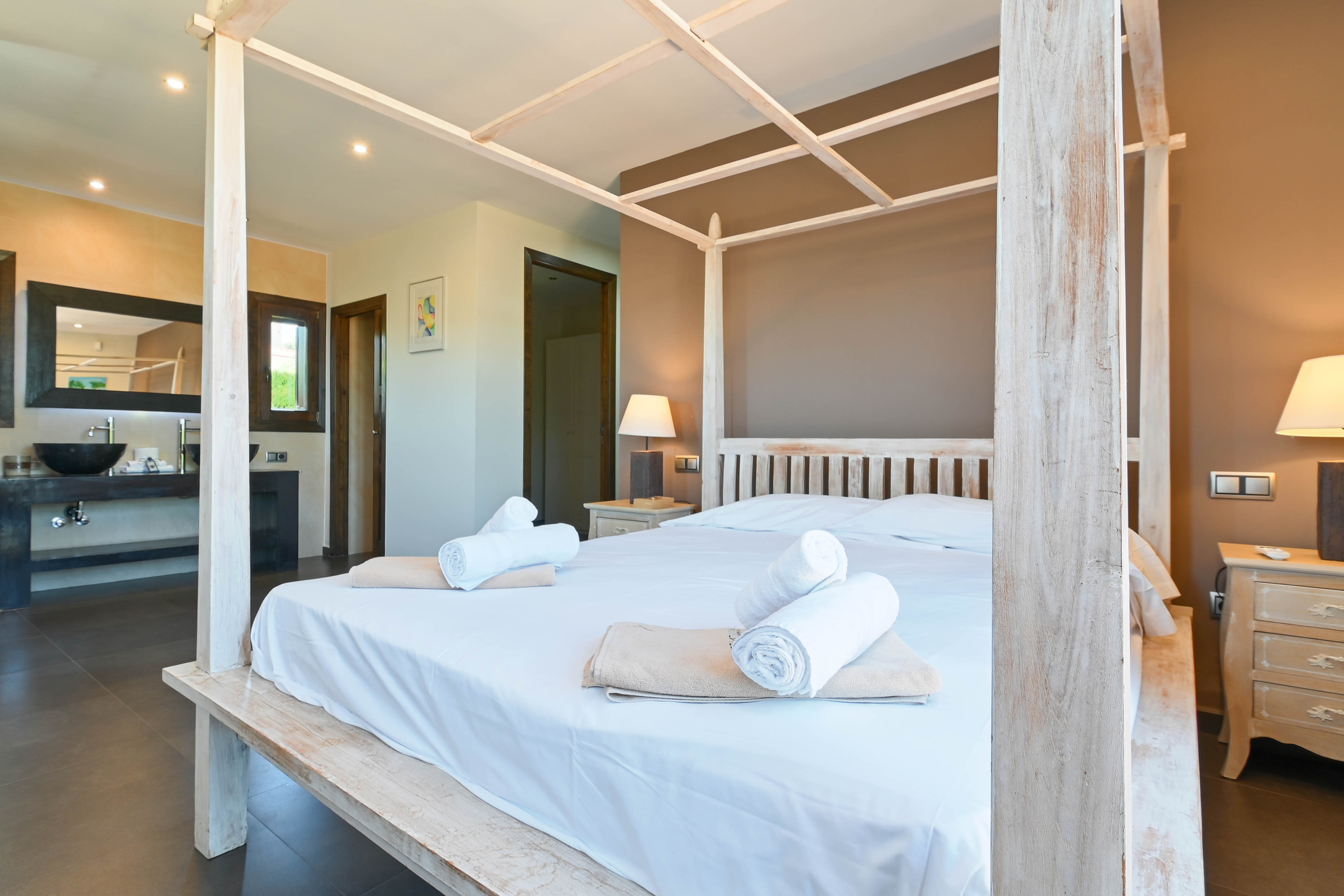 Bedroom with a white wooden canopy bed, rolled towels, and bathroom with a modern sink.