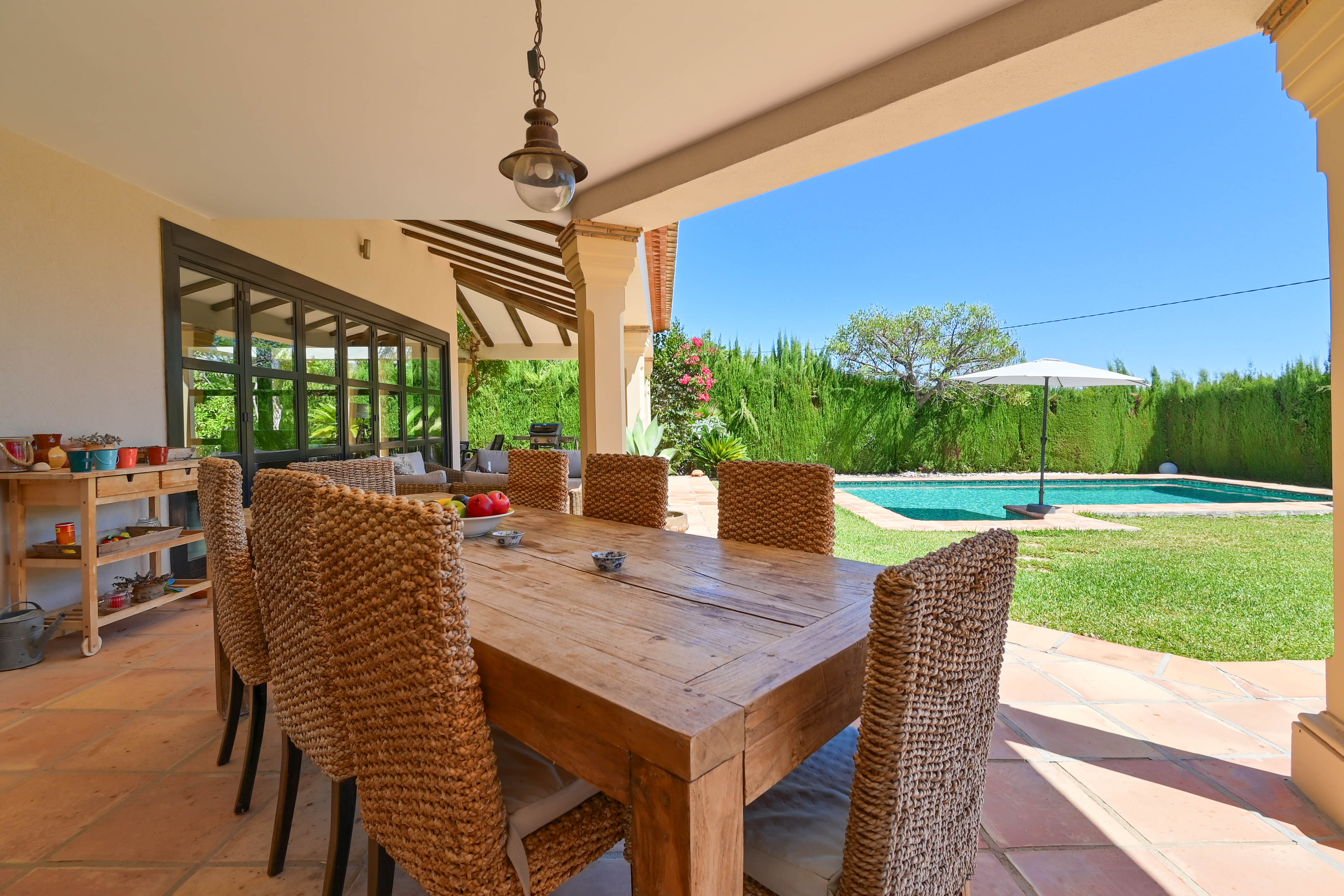 Covered outdoor dining area with solid wood table and rattan chairs overlooking the pool and garden.