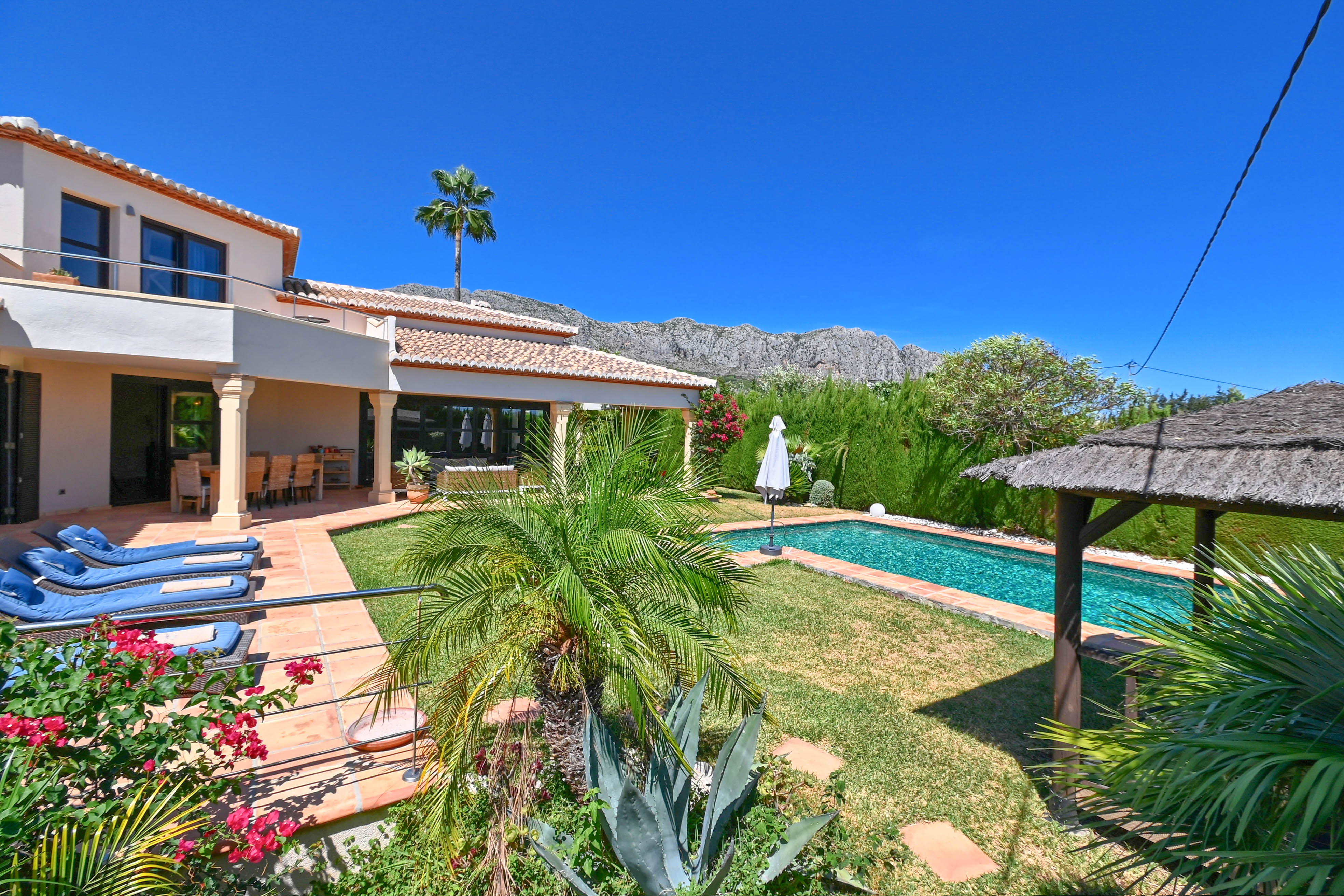 Garden view with rectangular pool and thatched gazebo, surrounded by lush greenery and mountains.