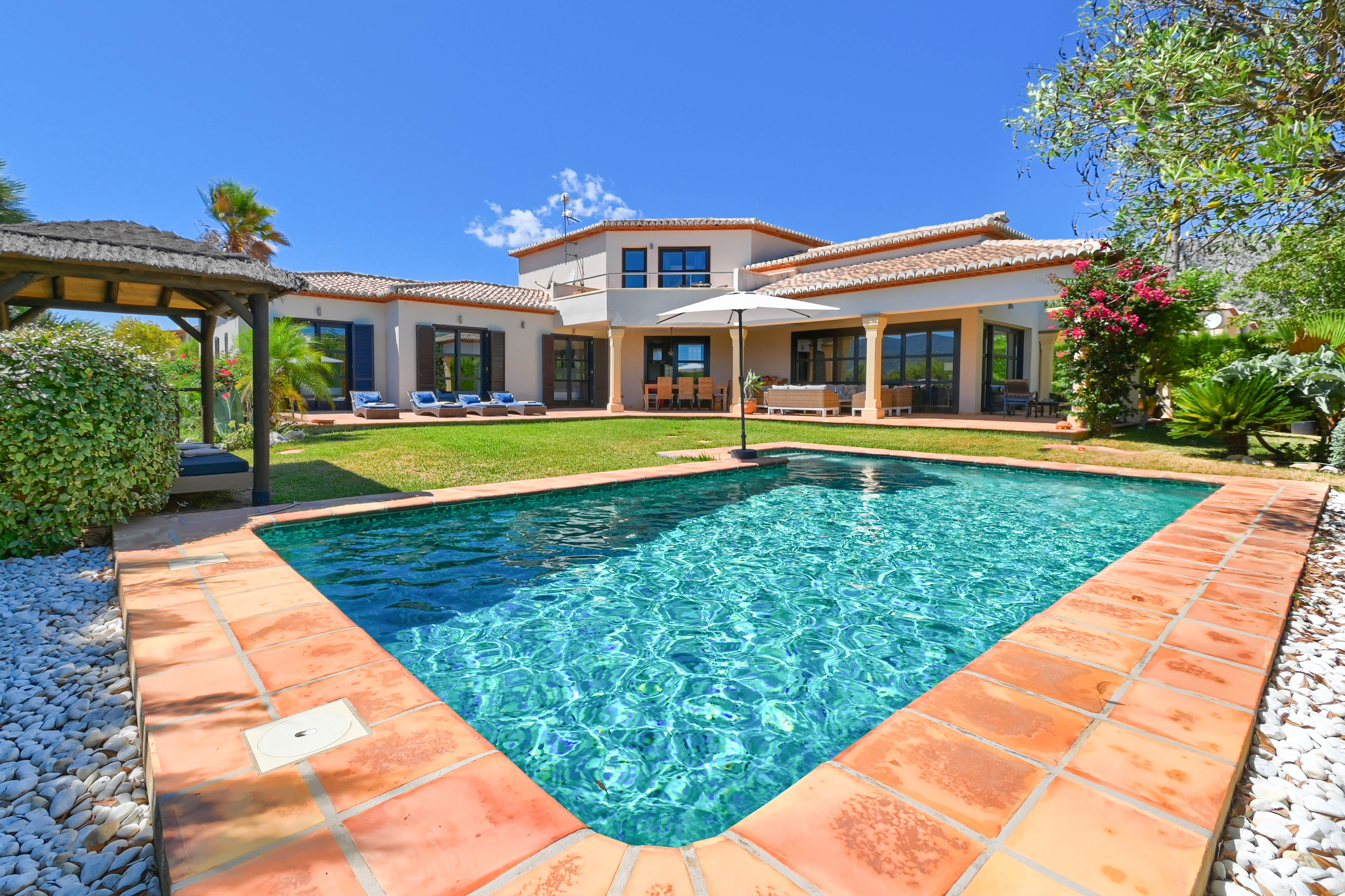 View of the turquoise pool with terracotta edging and the villa with garden and gazebo.