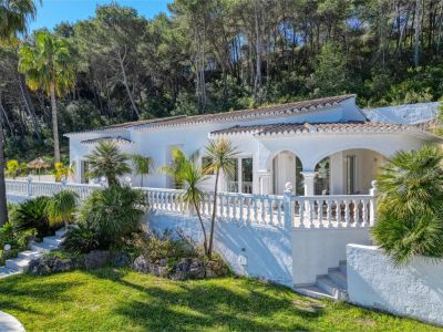 Exterior view of the villa featuring a covered terrace, balustrade railing, and a garden with Mediterranean greenery.