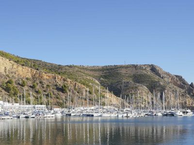 View of the marina with multiple boats docked and reflections in the water, with rocky mountains in the background.