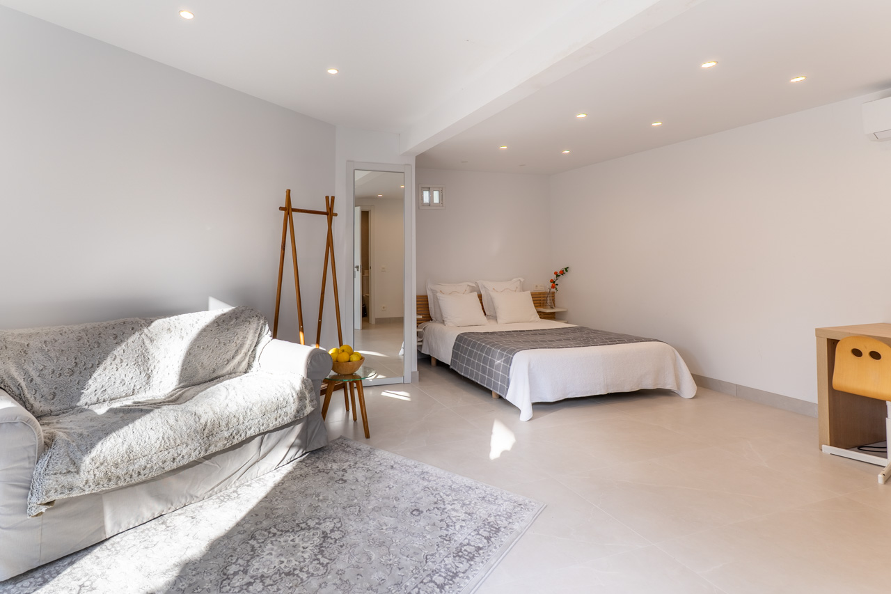 Bright bedroom with a sofa, double bed, and desk, illuminated by recessed spotlights.