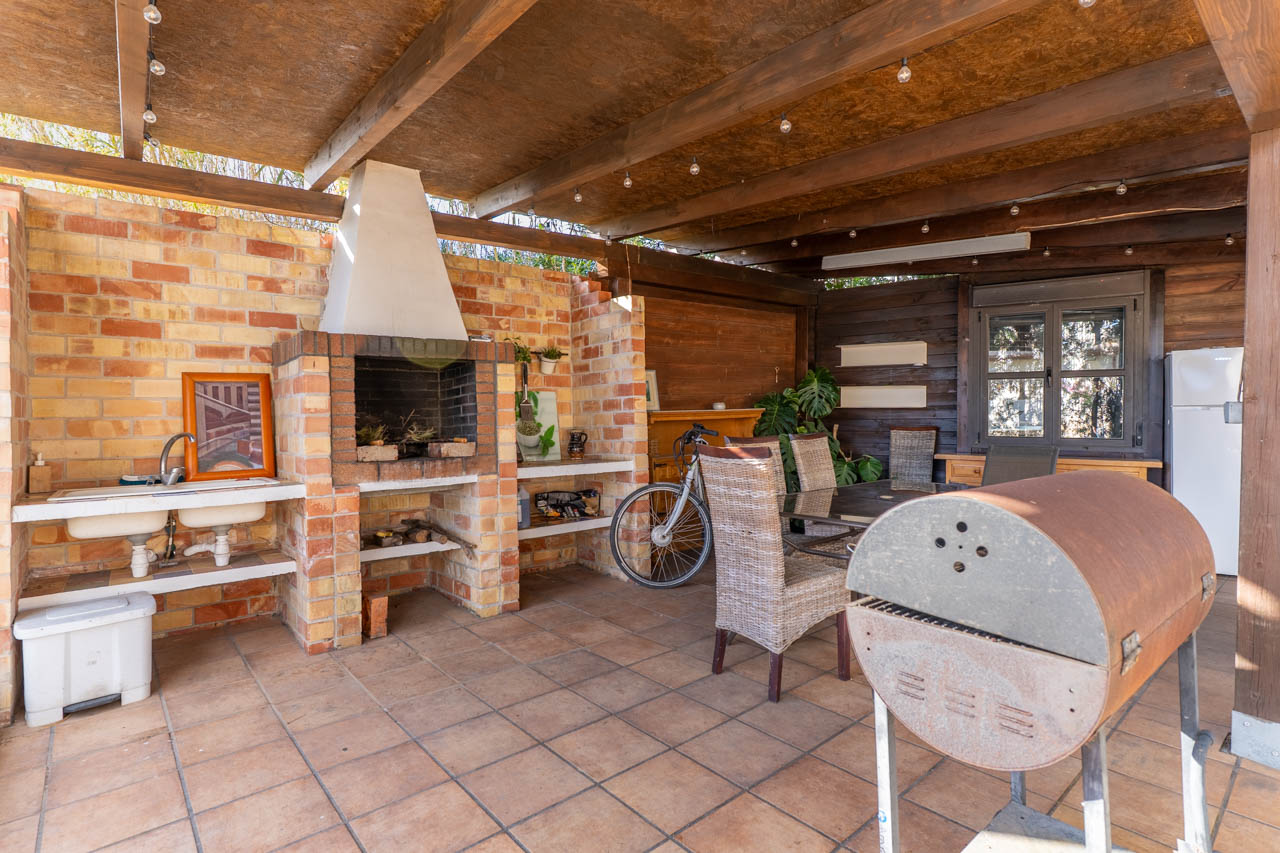 Outdoor barbecue area featuring a brick oven, double sink, and dining space under a wooden roof with ambient lighting.