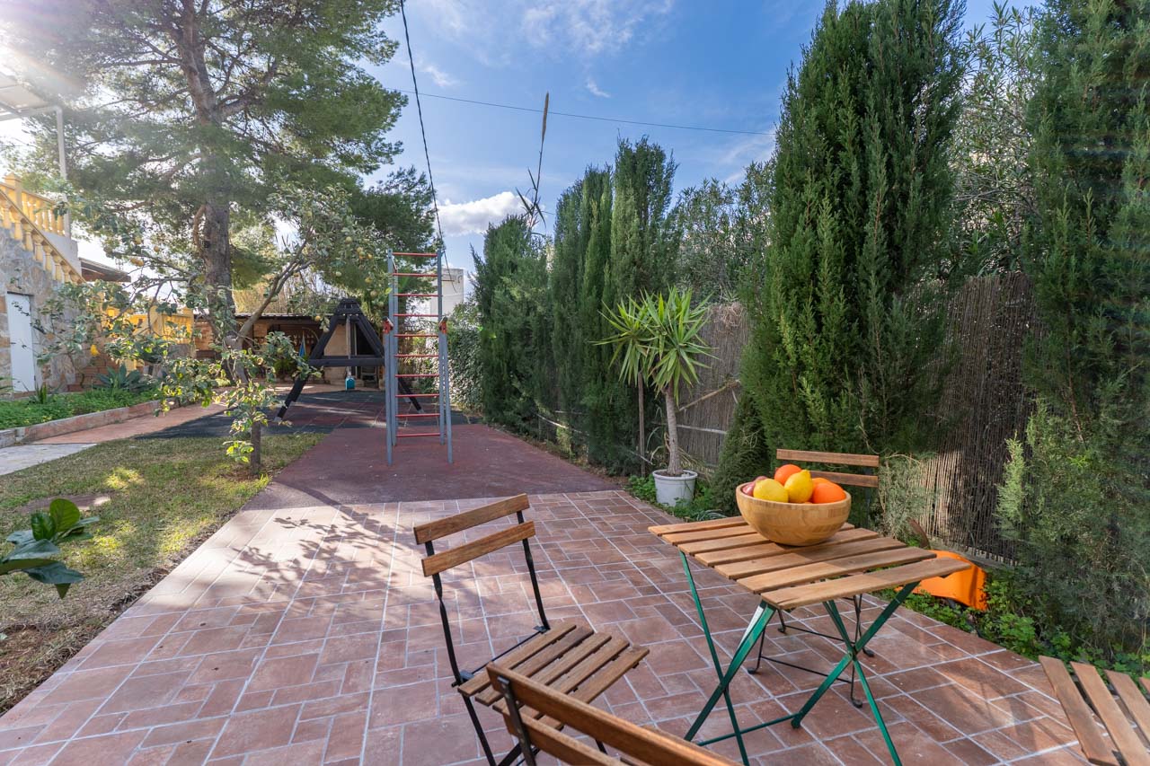 Terrace with wooden table and fruit bowl, surrounded by greenery and a children's play area.