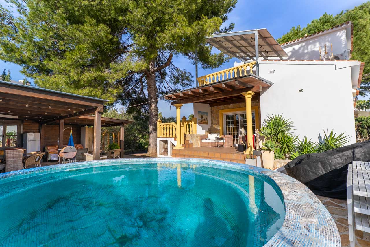 View of the pool with blue mosaic tiles and the covered terrace with yellow pillars.