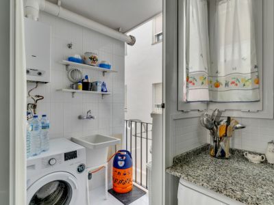 Laundry area featuring a washing machine, utility sink, and white tiled shelving. Window with patterned curtains.