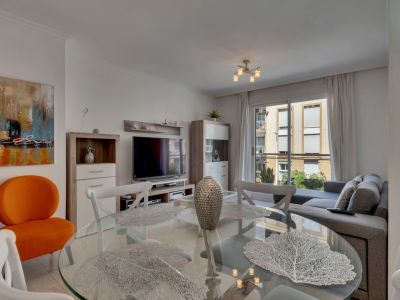 Living-dining room featuring a glass table, white chairs, and a gray sofa, lit by natural light.