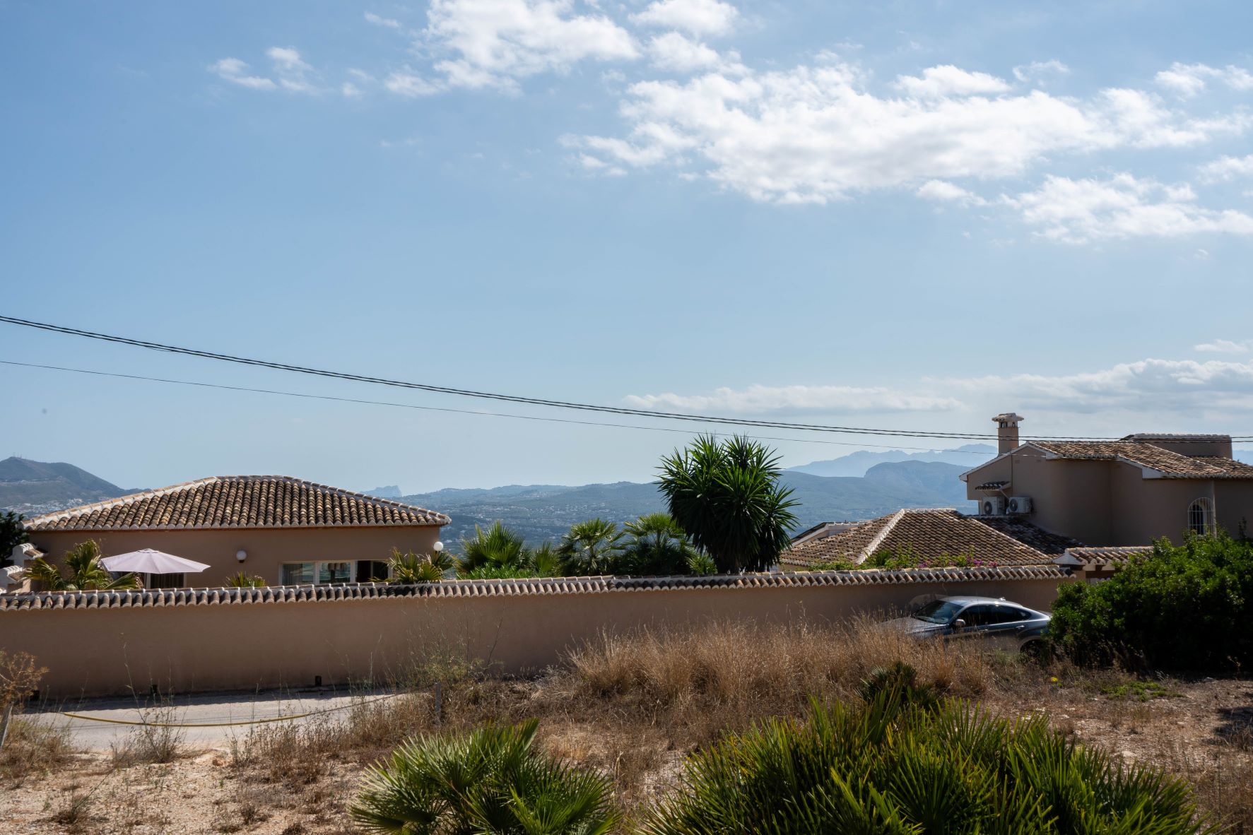 View of the plot with neighboring houses, vegetation and mountains in the background under a partly cloudy sky.