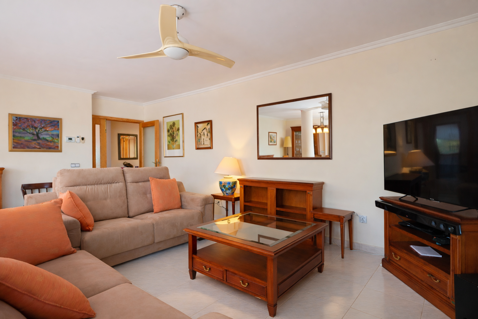 Furnished living room with beige sofa, wood coffee table, and TV, illuminated with natural light and lamp.