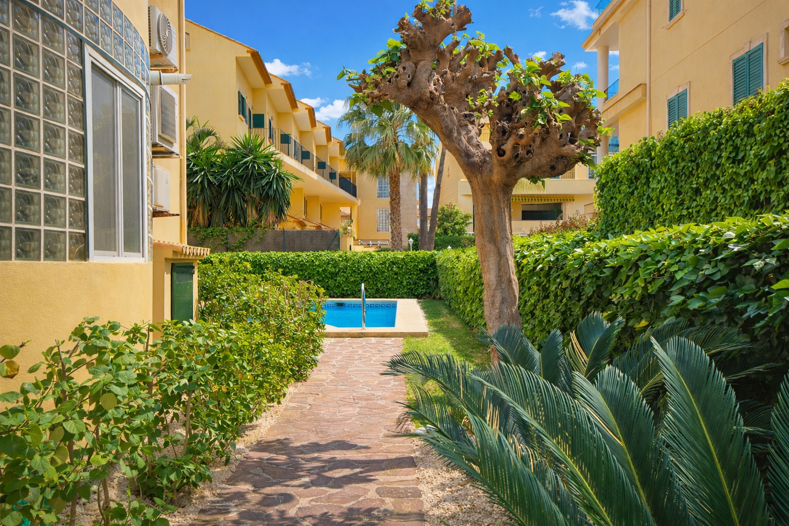 Stone path leading to a pool, surrounded by gardens and yellow buildings.