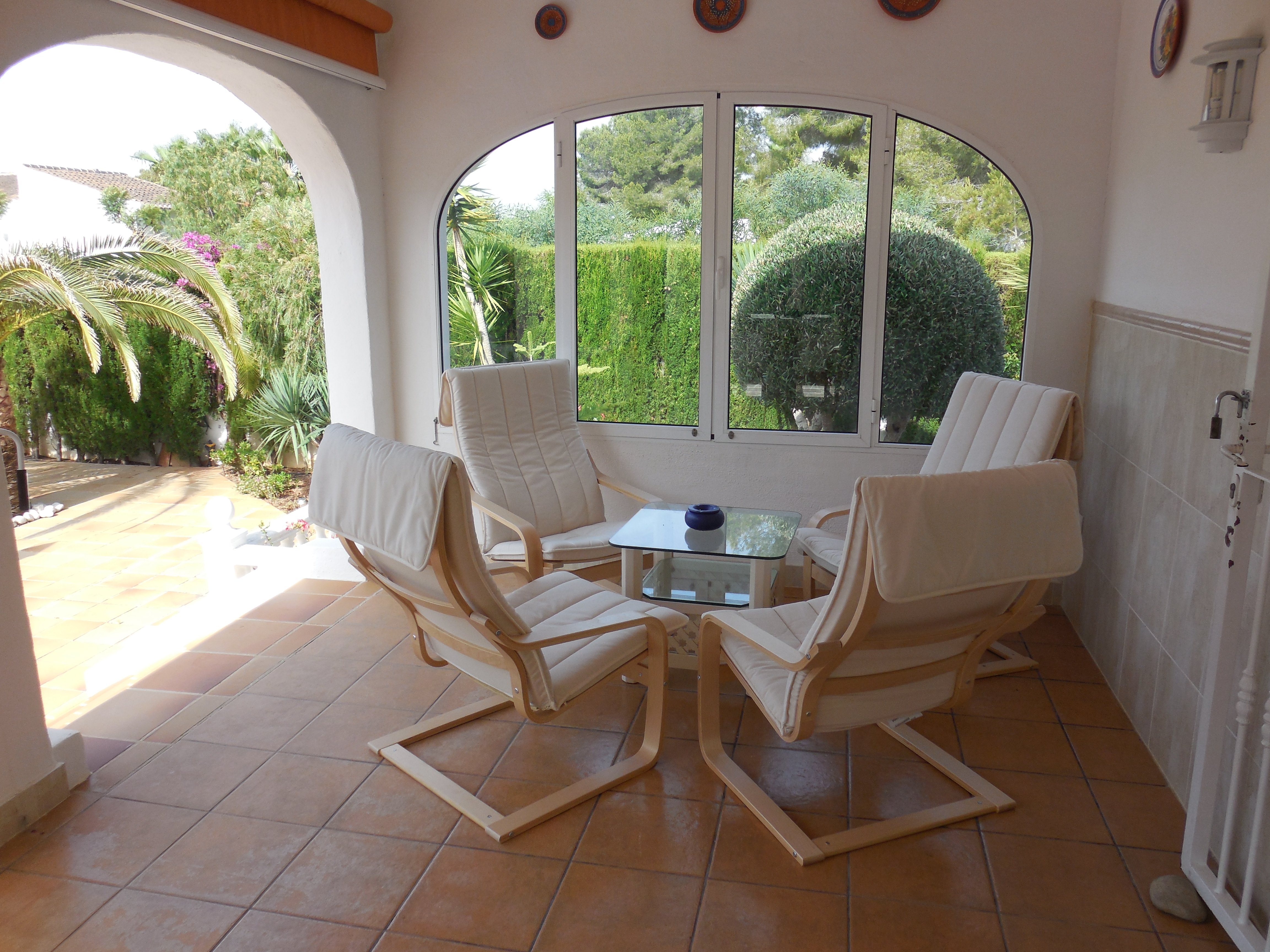 Covered terrace featuring four light wood armchairs with white cushions, a glass table, and garden views.