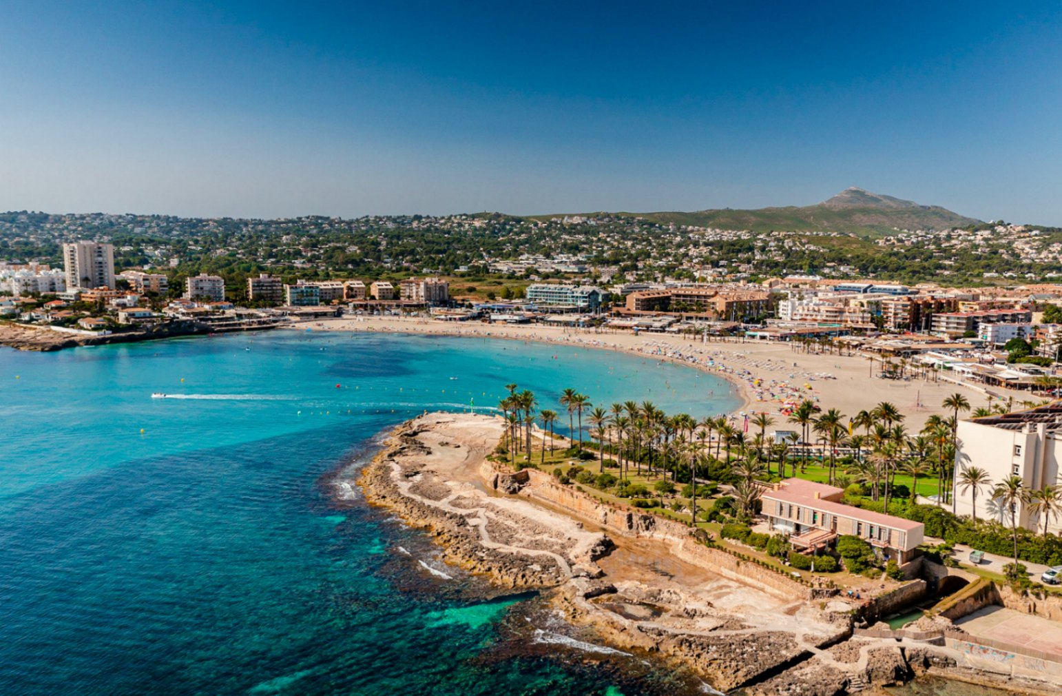 Aerial view of Playa El Arenal in Jávea, featuring its rocky coastline and surrounding buildings under a clear sky.