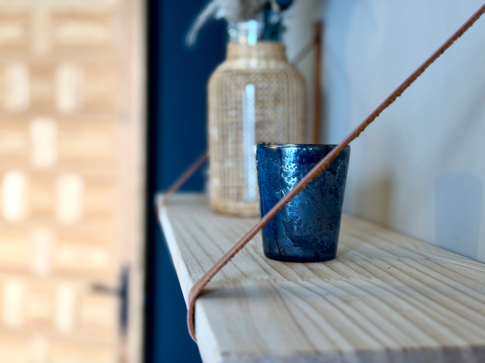 Close-up of a wooden shelf with a decorative blue glass and a wicker bottle in the background.