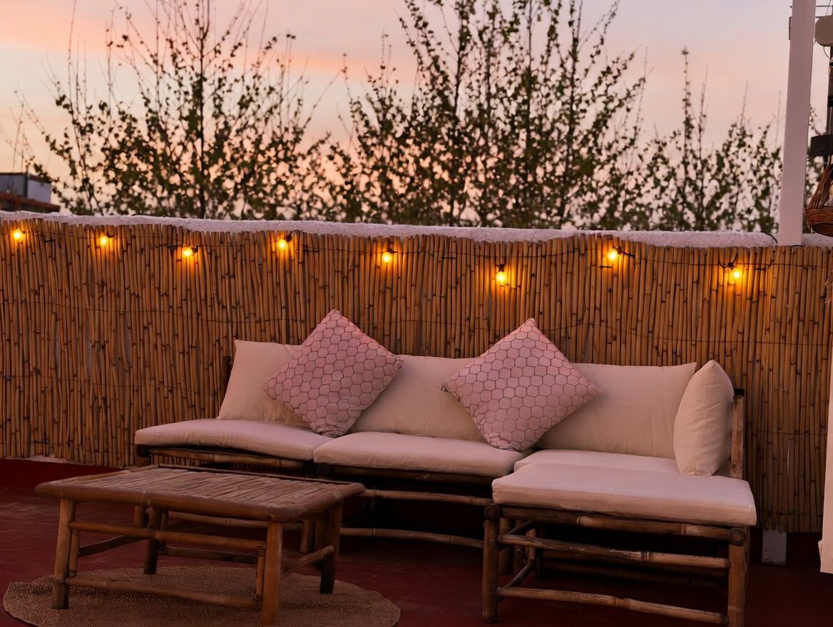 Terrace featuring a bamboo sofa with patterned cushions, illuminated by warm string lights, with a sunset sky view.