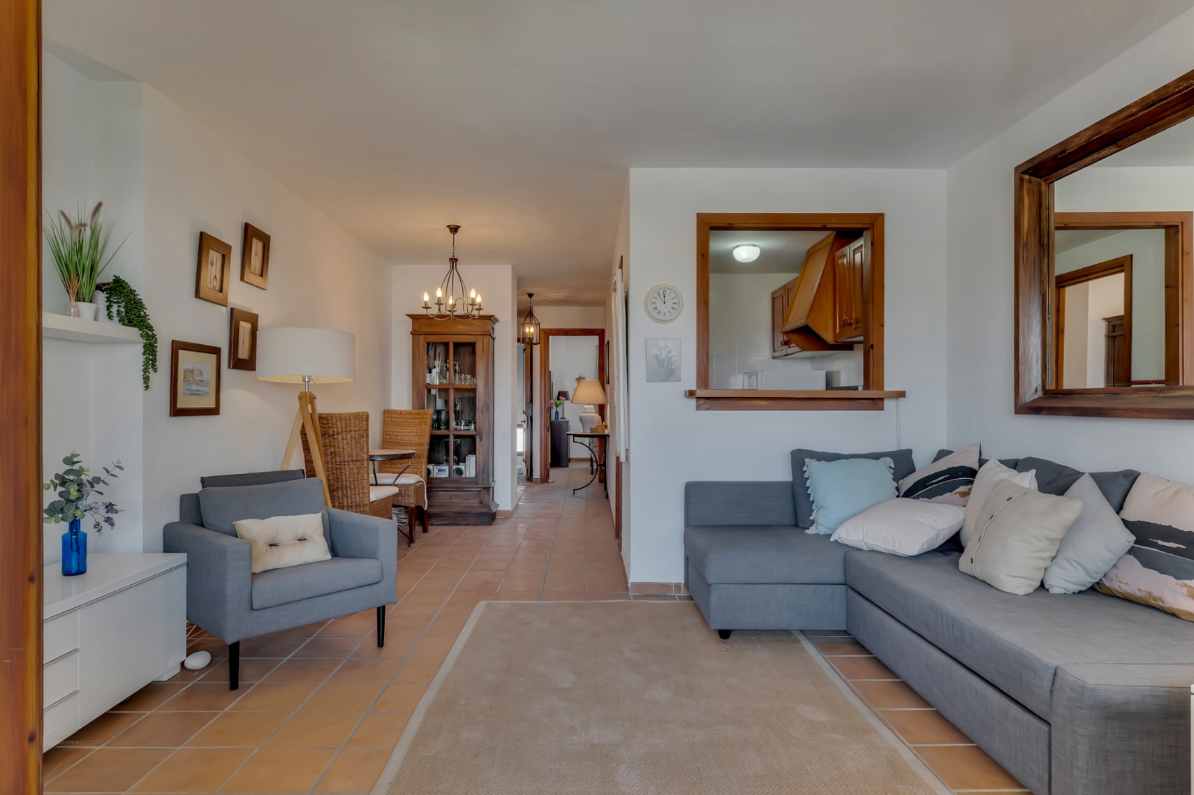 Living room with gray sofa, wood-framed mirror, and a view into the kitchen through an interior window.