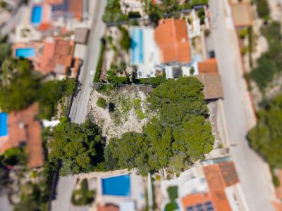 Aerial view of an undeveloped plot with trees, bordered by roads and residential properties featuring private pools and red roofs.