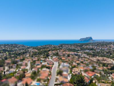Panoramic aerial view of the Benissa coast, featuring the Mediterranean Sea, the Peñón de Ifach, and a residential area with lush vegetation under a clear blue sky.
