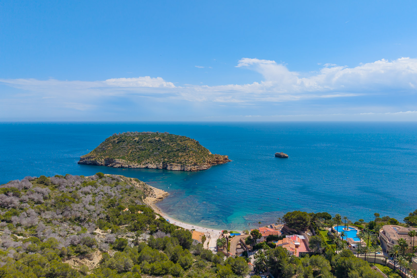 Aerial view of Jávea's coastline featuring Isla del Descubridor, beach, and residential area with a pool.