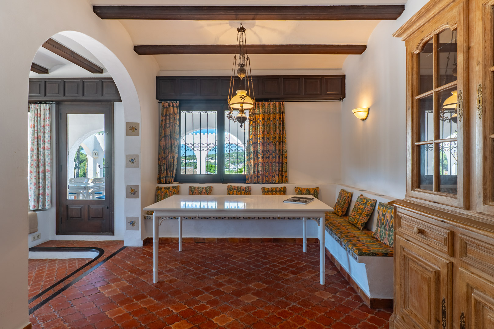 Dining room featuring a built-in bench, white table, and terracotta tile floor, illuminated by a chandelier.