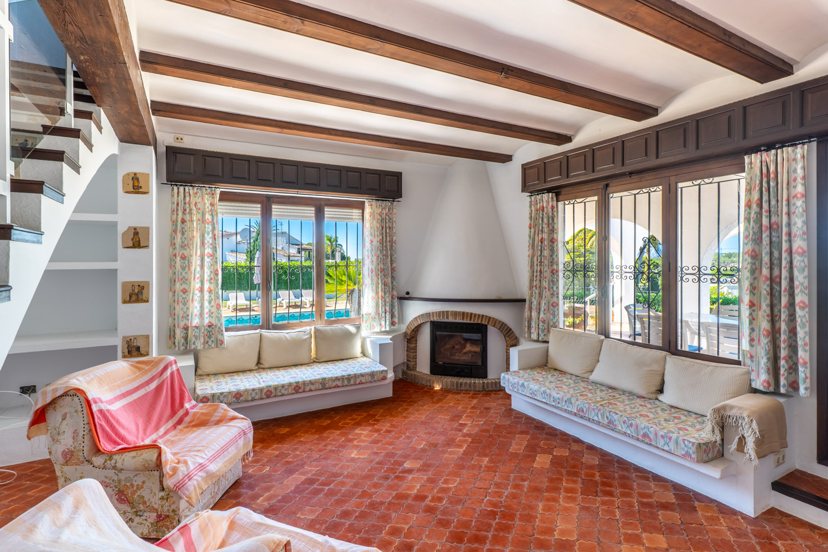 Living room featuring terracotta tile flooring, wood beams, and windows overlooking the pool.