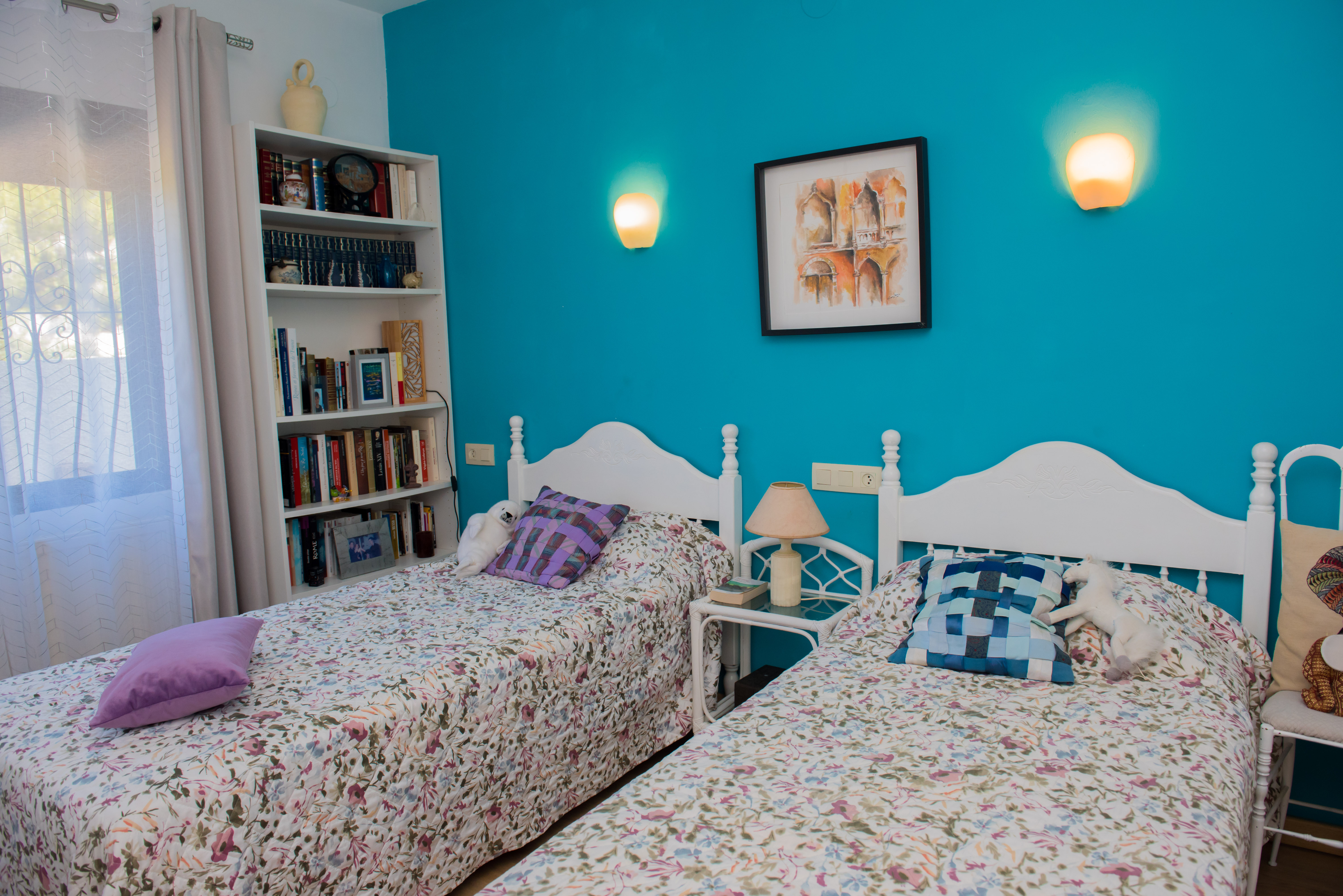 Bedroom with twin beds, turquoise wall, and a white bookshelf filled with books.